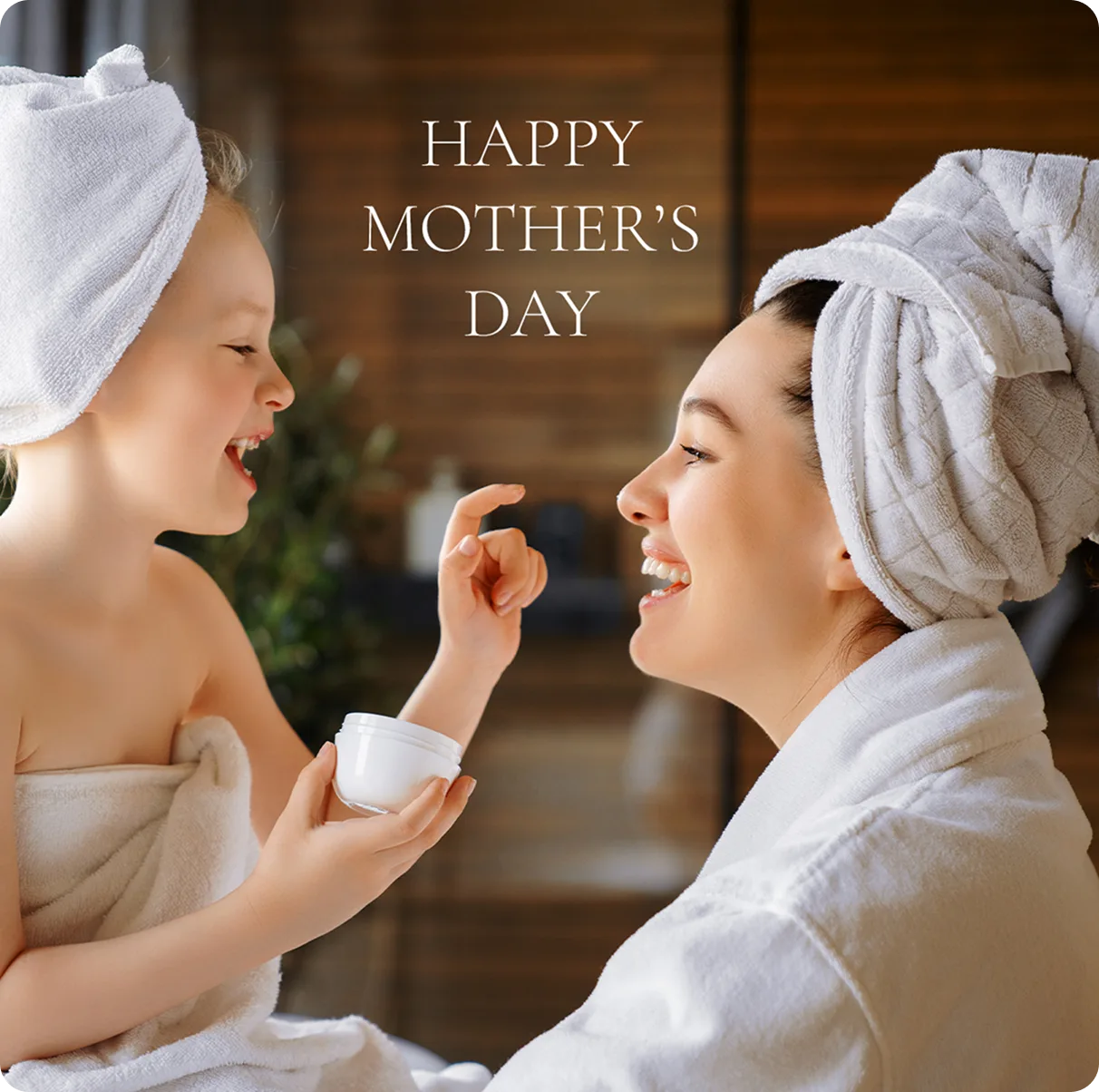 Happy Mother's Day image showing a mother and her young daughter in spa towels, laughing together as the daughter playfully applies skincare cream to her mom's nose